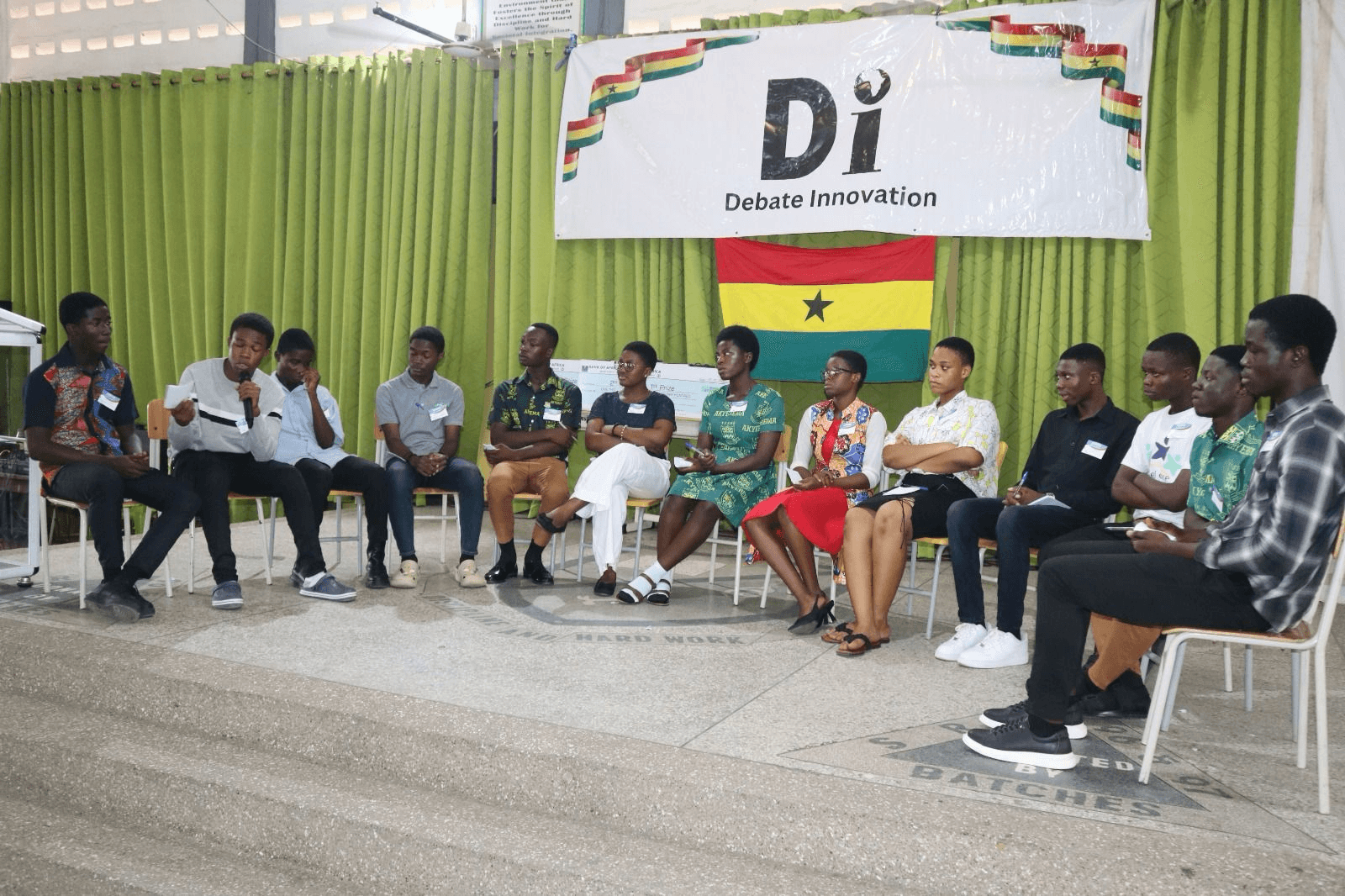 Senior high students seated in a semicircle mid-debate, beneath the Debate Innovation banner and the Ghana flag